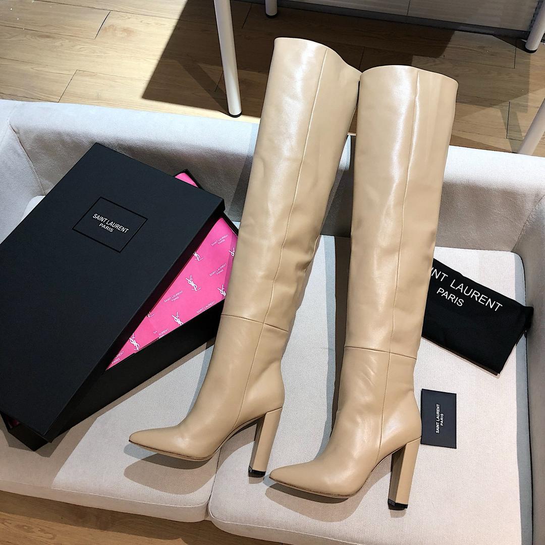 Beige knee-high boots with Saint Laurent packaging on a white surface.